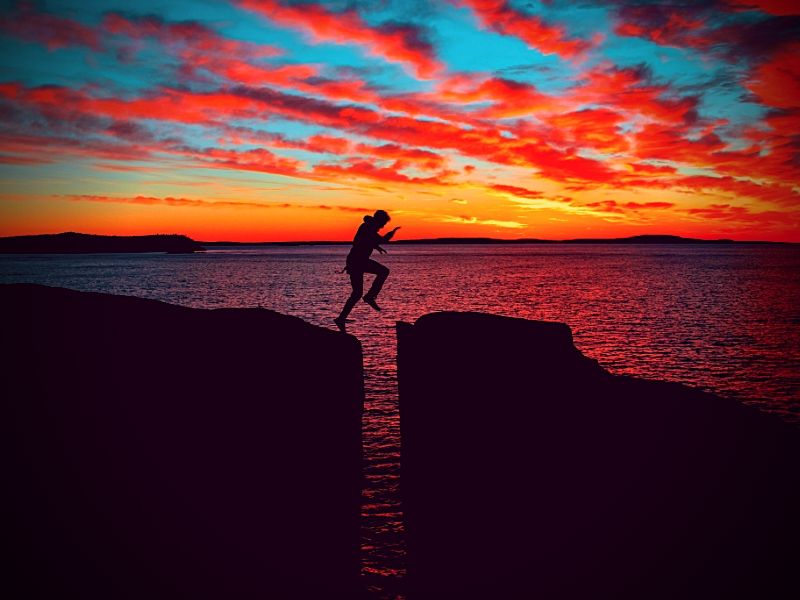 person jumping a chasm by the ocean in front of a bright sky